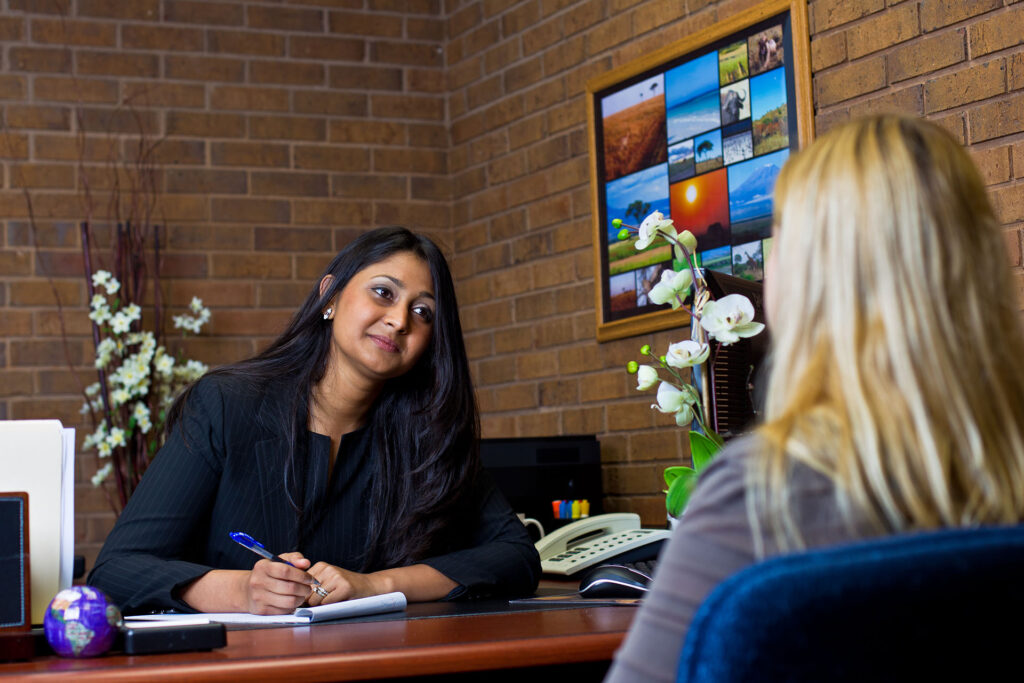 Professional woman attentively listening and taking notes while speaking to another person across the desk in an office setting.