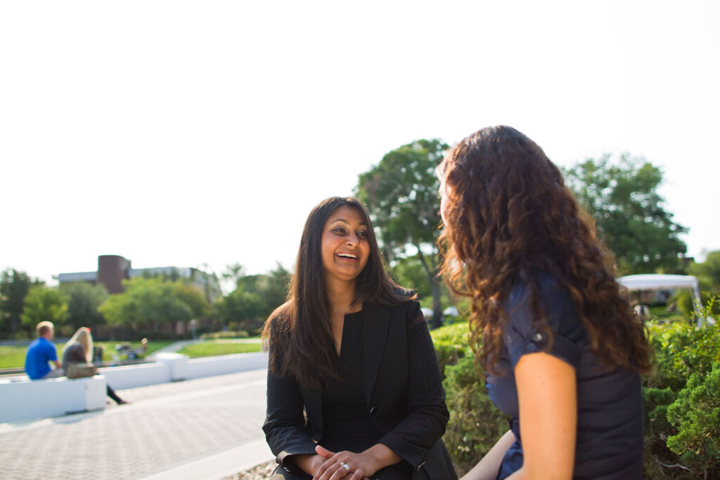 Professional woman smiling during a relaxed outdoor conversation with another woman, with trees and open campus space behind them.